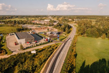 Valmiera, Latvia - August 8, 2024 - Aerial view of a gas station, trucks, and buildings near a highway in a rural landscape with greenery and distant structures. clipart