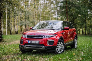 Valmiera, Letonya - 4 Ekim 2024 - Kırmızı bir Range Rover, arka planda ağaçlar olan ormandaki yeşil çimlere park etmiş..