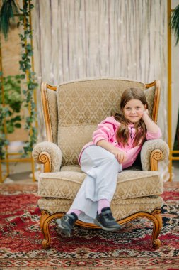 A young girl sits comfortably in an ornate armchair, wearing a pink hoodie and relaxed expression, against a decorative background with drapes and greenery. clipart