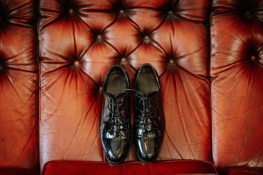 Valmiera, Latvia - August 24, 2024 - A pair of polished black dress shoes is displayed on a red, tufted leather sofa, creating a contrast of textures and colors. clipart