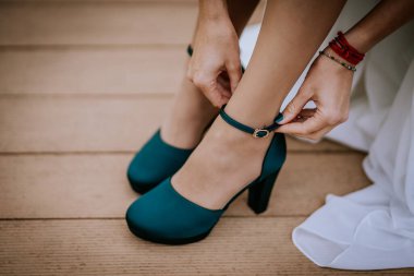 Valmiera, Letonya - 24 Ağustos 2024 - Yüksek topuklu ayakkabısının bilek kayışını bağlayan bir kadının yakın çekimi, ahşap bir güvertede nikah giysisine son bir dokunuş ekliyor..