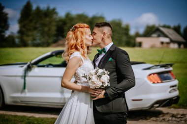Valmiera, Letonya - 24 Ağustos 2024 Gelin ve damat, beyaz bir spor arabanın önünde bir buket çiçek ve manzaralı bir kırsal arka plan ile öpüşüyorlar..