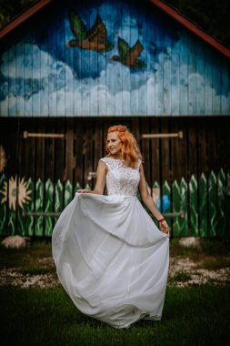 Valmiera, Letonya - 24 Ağustos 2024 - Kırsal bir düğün gününde yeşil kuşlarla çevrili renkli bir ahır duvarının önünde beyaz elbiseli, kızıl saçlı gelin.
