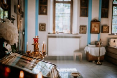 Valmiera, Latvia - August 24, 2024 - The image shows a lit candelabrum with a red glass candle holder in an Orthodox church, with religious icons and soft sunlight filtering through the window. clipart