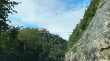 Reutte, Avusturya - 15 Eylül 2024 - Parçalı bulutlu bir gökyüzünün altında, yemyeşil ormanlarla çevrili bir tepedeki bir kaleye bağlanan asma köprünün manzaralı bir görüntüsü. Highline 179