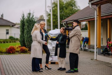 Valmiera, Letonya - 10 Ekim 2024 - Bir kameraman olay yerini kaydederken, elinde mikrofon olan bir muhabir iki kadınla dışarıda röportaj yapıyor. Arkaplanda yeşillik ve binalar yer alıyor.