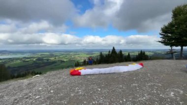 Bavyera, Almanya - 15 Eylül 2024 - Paraglider, parçalı bulutlu bir gökyüzü altında geniş yeşil bir vadiye bakan renkli bir paraşütle dağ yamacından havalanıyor..