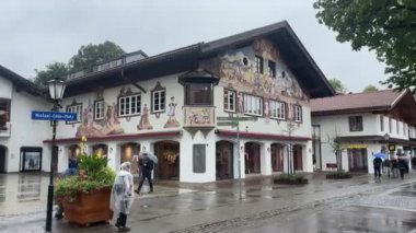 Bavyera, Almanya - 15 Eylül 2024 - Yağmurluk giymiş insanların yürüdüğü yağmurlu bir kasaba meydanında cephede boyalı fresklerle süslü bir bina.