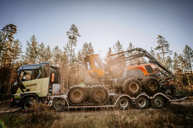 Gulbene, Letonya - 15 Ekim 2024 - Kamyon, ormandaki bir karavanda büyük ağaç kesme ekipmanları taşıyor, güneş ışığı ağaçların arasından gözetliyor, endüstriyel ormancılık çalışmaları yapıyor..