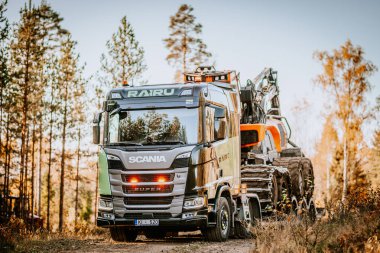 Gulbene, Letonya - 15 Ekim 2024 - Parlak bir gökyüzü altında sonbahar ağaçlarıyla çevrili, kereste makineleri taşıyan büyük bir kamyon, endüstriyel güç aktarıyor..