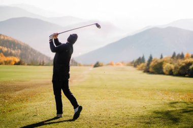 Golf sahasında sallanan bir golfçü, arka planda dağlarla ve yumuşak güneş ışığı altında sonbahar ağaçlarıyla manzaralı bir golf sahasında..