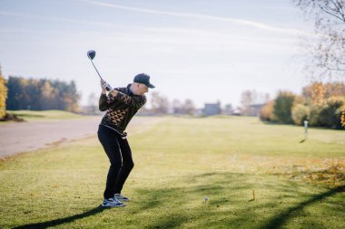 Güneşli bir günde golf sahasında sallanan bir erkek golfçü çimenli bir yeşilde topa vurmaya hazırlanıyor..