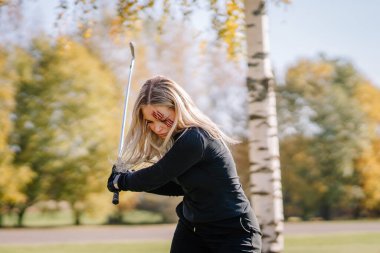 Yüzünde takma pençe izleri olan bir kadın, yoğun bir şekilde golf salıncağına odaklanmış, güneşli bir günde açık havada, Cadılar Bayramı temasına uygun..