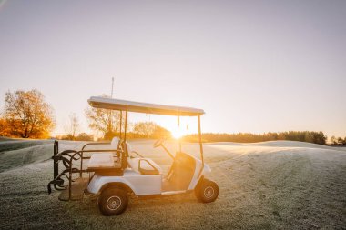 Valmiera, Letonya - 18 Ekim 2024 - Güneşin doğuşunda soğuk bir golf sahasına park etmiş bir golf arabası, çimenli arazide uzun gölgeler bırakan altın bir ışık..
