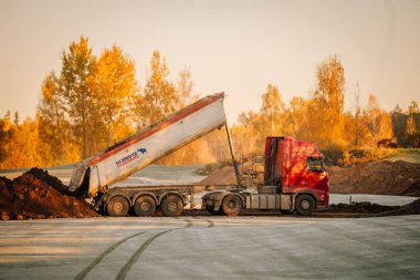 Valmiera, Letonya - 18 Ekim 2024 - Sonbahar ağaçları ve yumuşak sabah ışıklarıyla çevrili soğuk zemine toprak döken kırmızı bir kamyon.