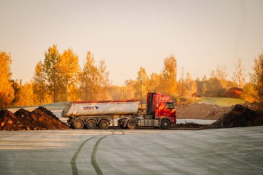 Valmiera, Letonya - 18 Ekim 2024 - Sabahın erken saatlerinde donuk zemine park etmiş, toprak ve sonbahar ağaçlarıyla çevrili kırmızı bir kamyon.