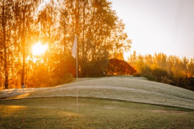Beyaz bir golf bayrağı, sonbahar ağaçlarıyla çevrili, altın gündoğumuyla aydınlanan, buzlu yeşil bir bayrağın üzerinde duruyor..