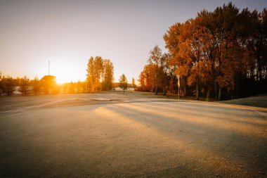 Güneşin doğuşuyla birlikte bir golf sahası, soğuk çimenler, uzun ağaç gölgeleri ve sonbahar yapraklarıyla çevrili bir bayrak direği..