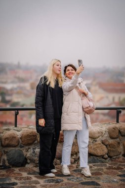 Vilnius, Lithaunia - October 22, 2024 - Two women posing for a selfie outdoors, standing on a stone platform with a cityscape blurred in the background. clipart