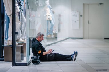 Valmiera, Letonya - 24 Ekim 2024 - Alışveriş merkezinde yerde oturan bir adam, yanında kamera olan bir telefonla, vitrin vitrininde mankenler olan bir adam.