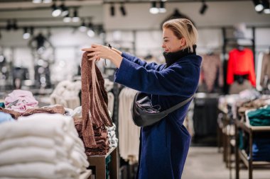 Valmiera, Latvia - October 24, 2024 - A woman in a blue coat is shopping in a clothing store, holding up a brown sweater, surrounded by folded garments and retail displays. clipart