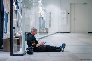 Alışveriş merkezinde yerde oturan bir adam, yanında kamera olan bir telefonla, vitrinin yanında mankenlerle oturuyor..