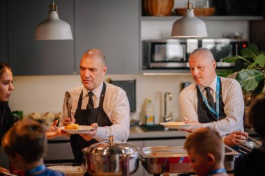 Riga, Latvia - October 25, 2024 - Two servers in formal attire serving food to guests in a buffet setting, with focused expressions in a modern kitchen environment. clipart