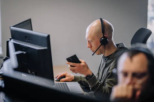 Riga, Letonya - 25 Ekim 2024 - Kulaklık takan bir adam modern bir ofiste akıllı telefon tutarken bilgisayar başında çalışır, çoklu görev ve iletişim üzerine odaklanır..