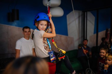 Riga, Latvia - October 25, 2024 - A young girl in a helmet enjoys a zipline activity during the Peter Pan Party, surrounded by friends and supervised by adults.