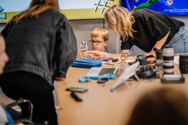 Riga, Letonya - 25 Ekim 2024 - Peter Pan Party 'de akıllı telefon kullanan gözlüklü genç bir çocuk, bir masanın üzerinde kamera ve cihazlarla çevrili bir şekilde yetişkinler tarafından yönlendiriliyor..