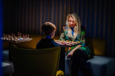 Riga, Latvia - October 25, 2024 - A woman and a boy engage in a thoughtful game of checkers at the Peter Pan Party, with chess pieces visible on a table in a softly lit room. clipart