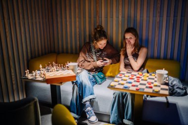 Riga, Letonya - 25 Ekim 2024 - İki kız rahat bir ortamda oturuyor, biri satranç ve dama tahtaları masada otururken, biri telefonu kontrol ediyor, PETER PAN Parti etkinliği sırasında.