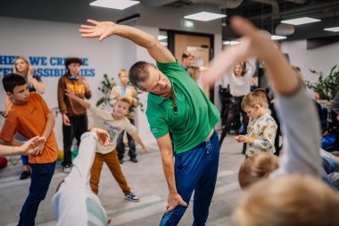 Riga, Letonya - 25 Ekim 2024 - Bir adam PETER PAN Partisinde çocuklara esneme egzersizinde liderlik ediyor, katılımcılar da onu eğlenceli ve enerjik bir iç mekan ortamında takip ediyor.