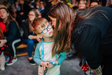 Riga, Letonya - 25 Ekim 2024 - Çocuk kalabalık bir odada dikkatle dinlerken bir kadın Peter Pan Partisi 'nde gözlük takan genç bir çocuğa fısıldıyor..