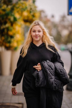 Vilnius, Lithaunia - 22 Ekim 2024 - Siyah kapüşonlu ve ceketli genç bir kadın arka planda sonbahar yapraklarıyla güvenle şehrin caddesinde yürüyor..