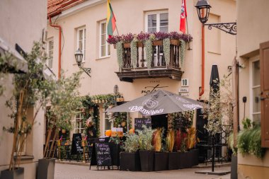 Vilnius, Lithaunia - October 22, 2024 - A charming outdoor cafe with flower decorations and patio seating under umbrellas on a cozy street corner, featuring a balcony and Lithuanian flags. clipart