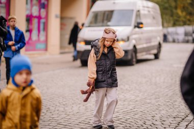 Vilnius, Lithaunia - 22 Ekim 2024 - kaldırım taşından bir sokakta oyuncak taşıyan genç bir kız, arkasında düşünceli bir ifade, insanlar ve bir minibüs bulunan kış kıyafeti giyiyor..