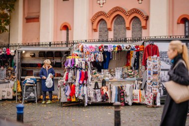 Vilnius, Lithaunia - 22 Ekim 2024 - Bir sokak marketi renkli kıyafetler, aksesuarlar ve hediyelik eşyalar satar. Yoldan geçen biri ön planda yürürken bir satıcı ögeleri düzenler.