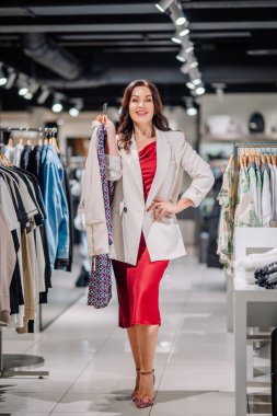 Valmiera, Latvia - October 24, 2024 - A confident woman in a white blazer and red dress shopping in a clothing store, holding a patterned jacket, with racks of clothes around her. clipart