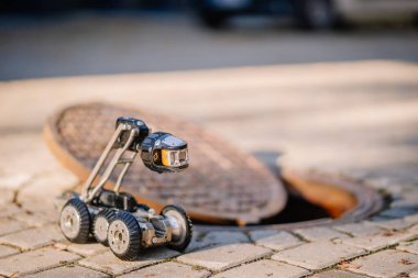 Tekerlekleri ve kamerası olan küçük bir teftiş robotu asfaltlı bir yüzeyde, açık bir rögar kapağının yanında, yeraltı keşfi için hazır bekliyor..