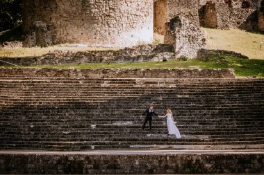 Valmiera, Letonya - 24 Ağustos 2024 Gelin ve damat tarihi harabelerin arkaplanına karşı antik taş basamaklarda el ele tutuşarak romantik ve zamansız bir sahne yarattılar..
