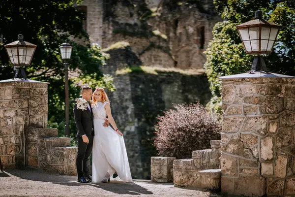 Valmiera, Letonya - 24 Ağustos 2024 - Gelin ve damat, yemyeşil ve antik mimari ile çevrili taş duvar ve fenerlerle poz vererek düğün günlerinde romantik bir an paylaşıyorlar..