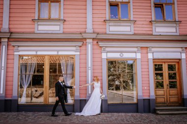 Valmiera, Letonya - 24 Ağustos 2024 Gelin ve damat klasik pembe bir binanın önünde el ele tutuşarak romantik poz veriyorlar. Gelin dalgalı beyaz bir elbise giyer, zamansız bir sahne yaratır..