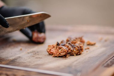 Valmiera, Letonya - 7 Kasım 2024 - Ahşap bir tahtada ızgara et dilimleyen eldivenli bir elin yakın çekimi, sulu dokuları ve kömürleşmiş kenarları gösteriyor kırsal bir barbekü sunumu için.