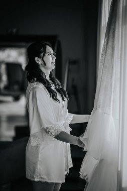 Koknese, Letonya - 26 Eylül 2023 - Dantel bornozlu bir gelin yumuşak ışıkta gelinliğine dikkatle bakar ve siyah beyaz renkte huzurlu ve samimi bir an yaratır..