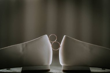 Zarif beyaz gelinlik ayakkabılarının topukları arasında dengelenmiş iki alyans yumuşak bir arkaplan karşısında sembolik ve minimalist bir kompozisyon oluşturuyor..