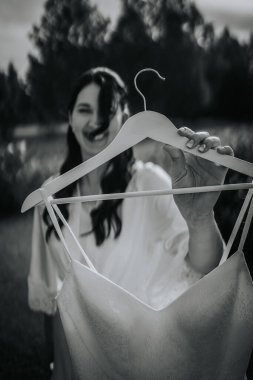 Koknese, Letonya - 26 Eylül 2023 - Bir gelin dantelli bir gelinlik ile ahşap bir askıya sarılmış, bulanık bir arka planda hafifçe gülümsüyor. Görüntü siyah ve beyaz, sonsuz bir his katıyor..