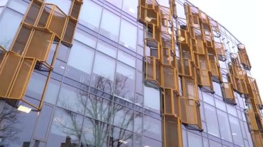 Cesis, Letonya - 13 Kasım 2024 - Ön cephesinde sarı metal paneller olan modern cam bina, pencerelerde çıplak ağaçların yansımaları olan eşsiz bir mimari tasarım yaratıyor..