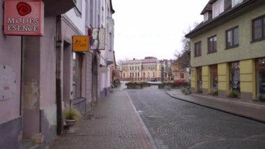 Cesis, Letonya - 13 Kasım 2024 - Eski bir kasabada Cobblestone Caddesi üzerinde dükkan tabelaları, renkli binalar ve bulutlu bir günde tarihi bir bina manzarası.