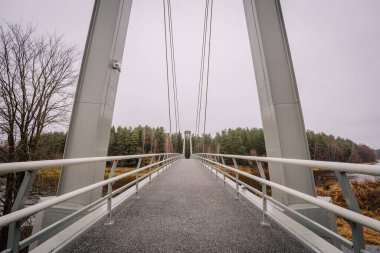 Valmiera, Letonya - 14 Kasım 2024 - Metal parmaklıklar ve kablolarla çevrili, bulutlu bir gökyüzünün altındaki ormanla çevrili modern bir asma köprü manzarası.